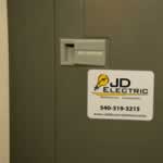 Yellow lightbulb and black text on white vinyl rectangle JD Electric weatherproof label on electrical box