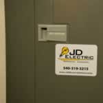 Yellow lightbulb and black text on white vinyl rectangle JD Electric weatherproof label on electrical box.