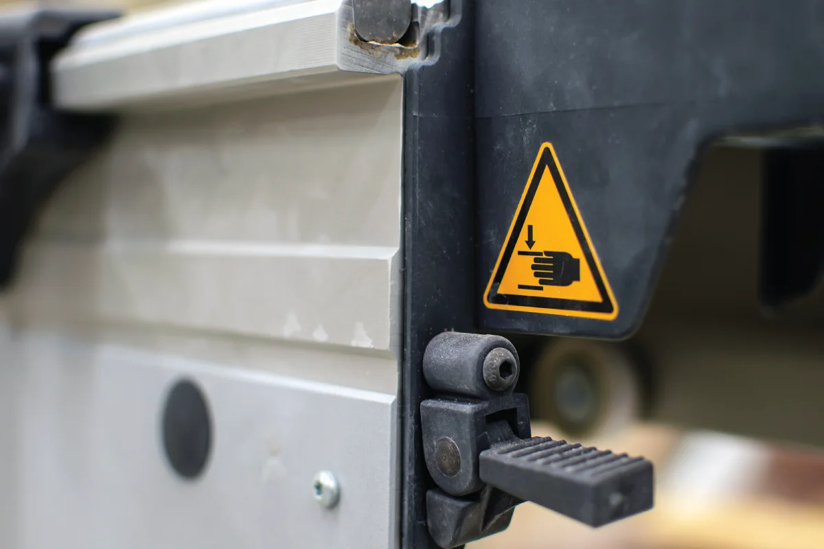 Equipment label showing a yellow triangle with a hand symbol and a downward arrow, warning of a potential pinch or crushing hazard, likely on a piece of machinery.